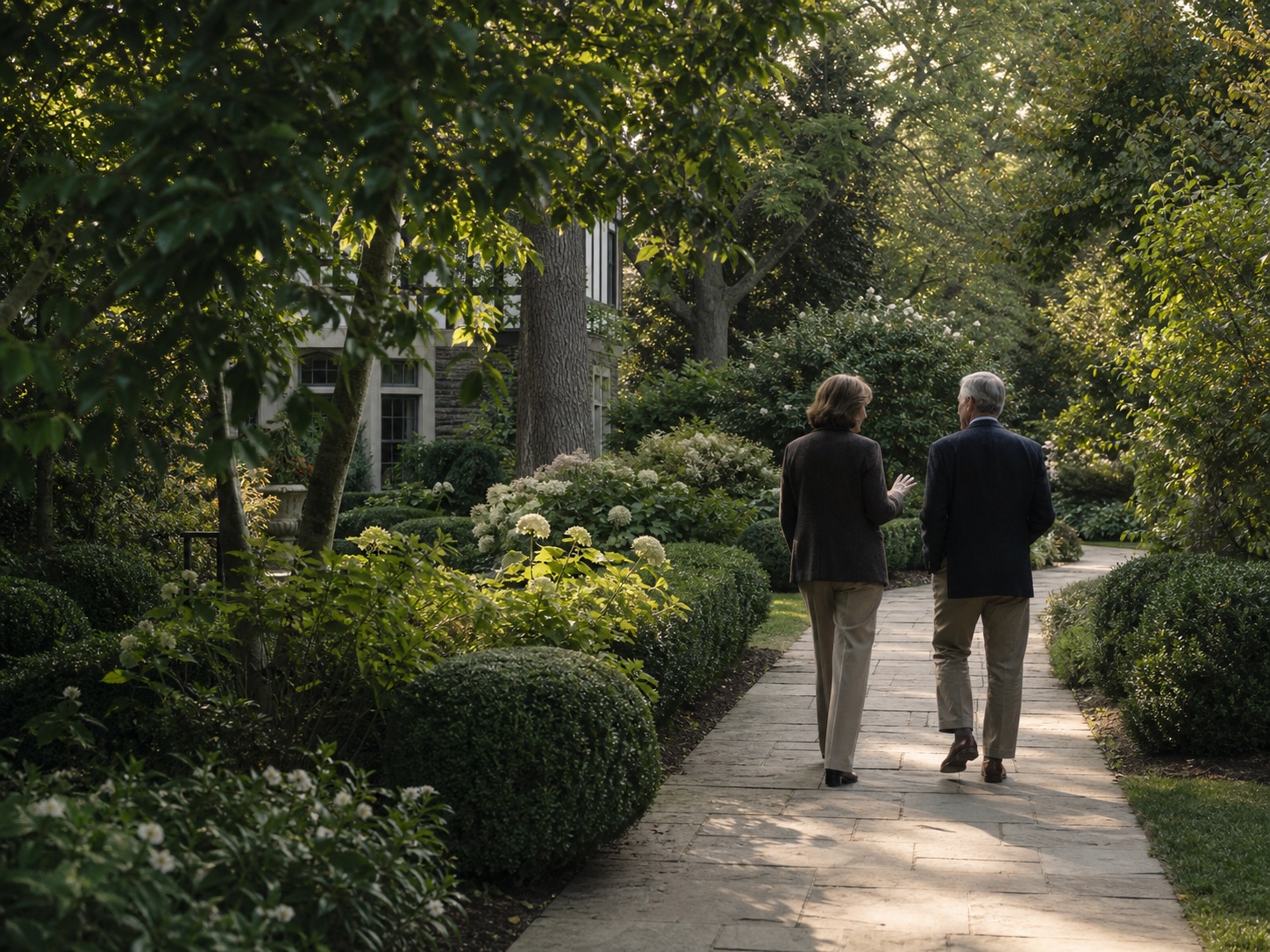 Two people walking along a garden path beneath mature trees
