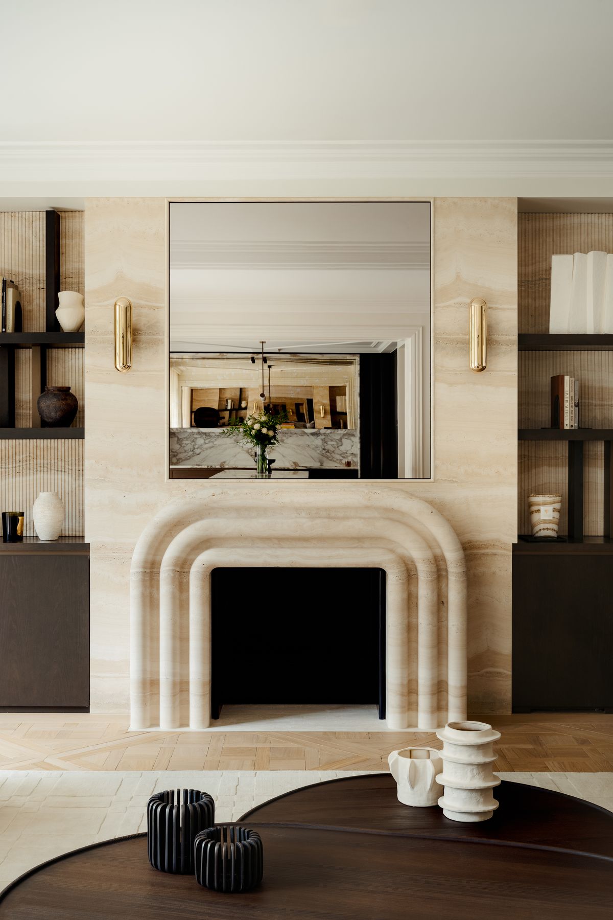 Illustrative pale fireplace surround beneath a large mirror, between dark shelves with books and decorative objects