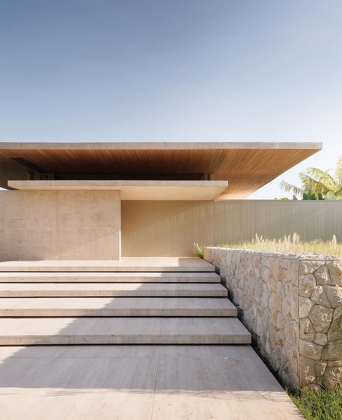 Wide pale steps beside a rough-textured wall lead toward an entry beneath a deep roof with a timber-toned underside.