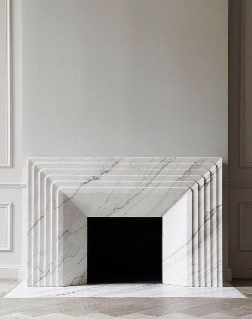 Pale veined fireplace surround with stepped rounded edges around a dark opening, against a paneled wall.