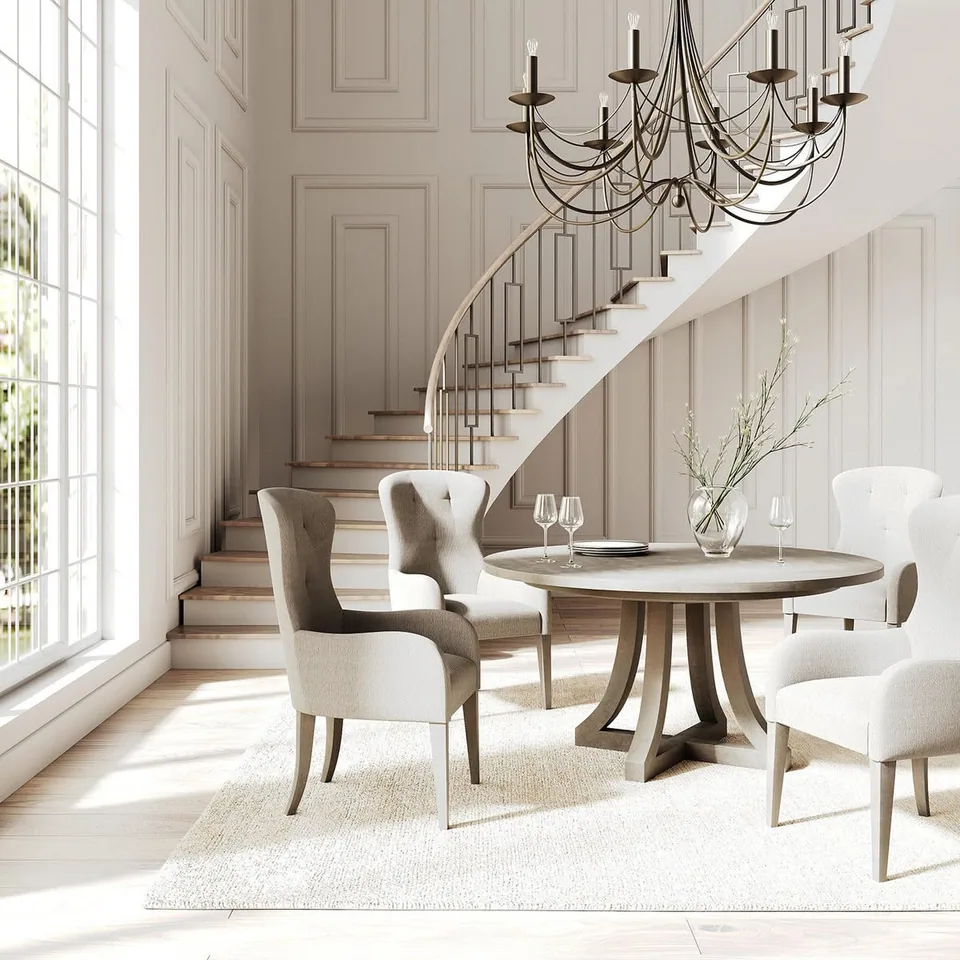 Bright pale dining room with a round table, upholstered chairs, tall multi-pane windows, a curved staircase, paneled walls, and a dark branching chandelier