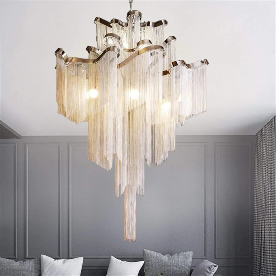 Illuminated tiered chandelier with reflective curved bands and hanging glass strands above a gray paneled wall and the top edge of a sofa