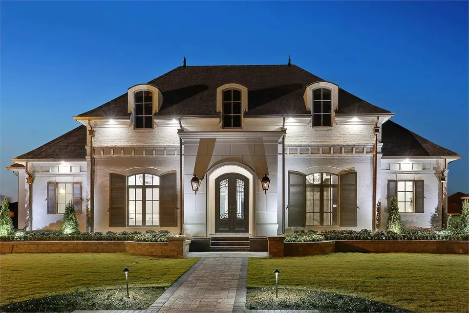 Frontal evening view of a symmetrical white residence with dormer windows, arched openings, facade lighting, and a straight walkway