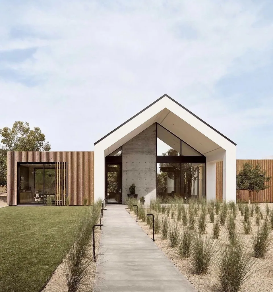 Gabled modern residence with a straight concrete walkway, ornamental grasses, wood screens, and full-height glazing