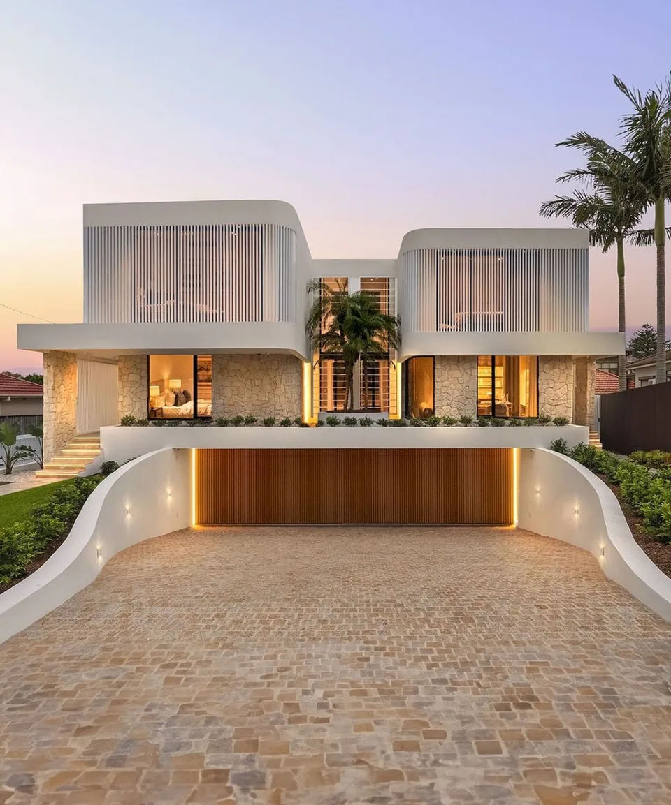 Front dusk view of a white two-story residence with vertical screens, pale block-patterned lower walls, palm trees, and a driveway descending to a brown slatted garage door