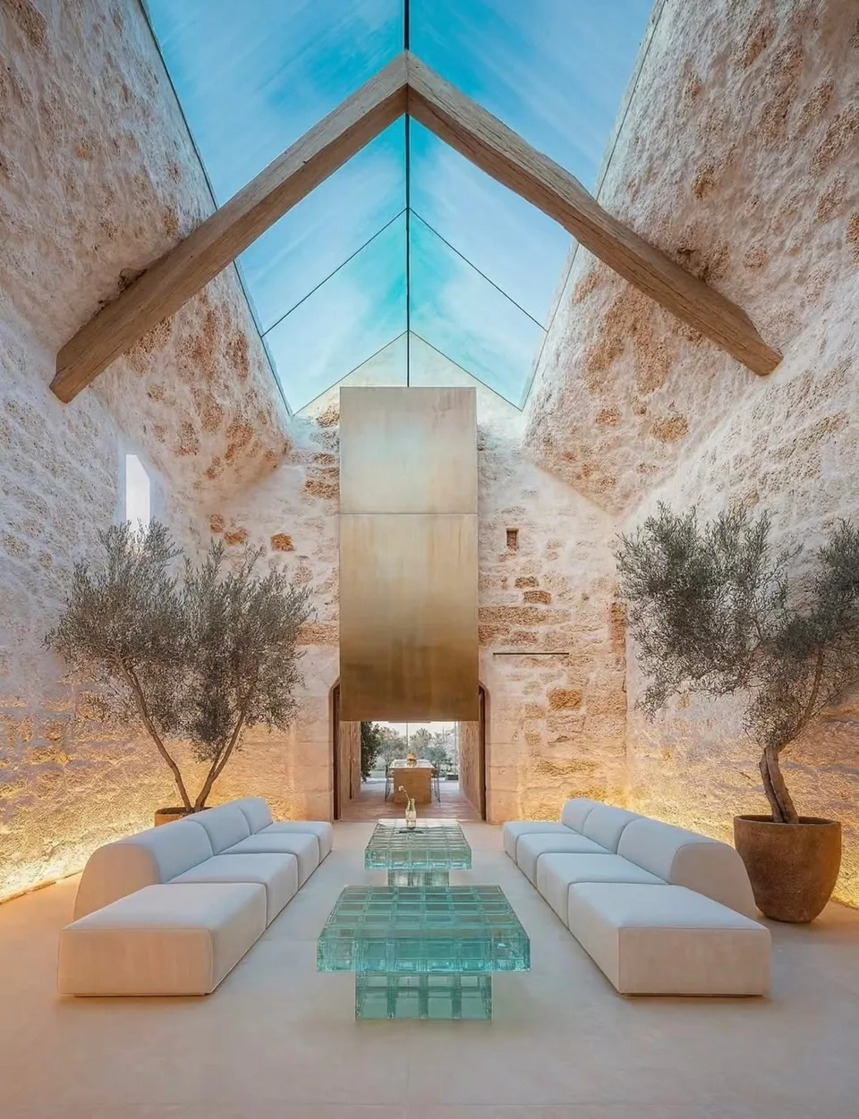 Stone-walled room with white sofas, glass block tables, two potted trees, and a glazed pitched roof