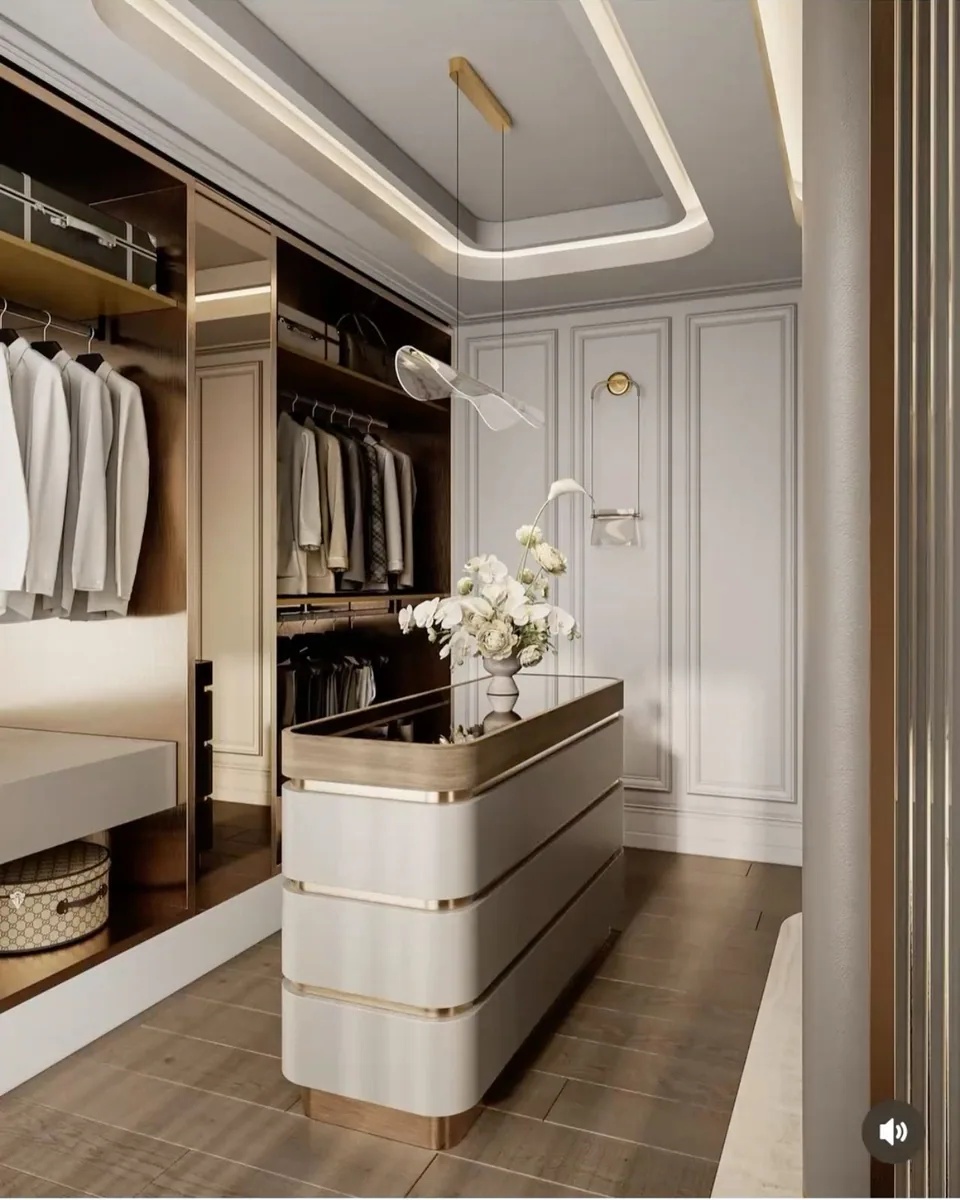 Pale walk-in closet with open hanging bays, an illuminated dark cabinet wall, a rounded central island, a low bench, and ribbed glass doors